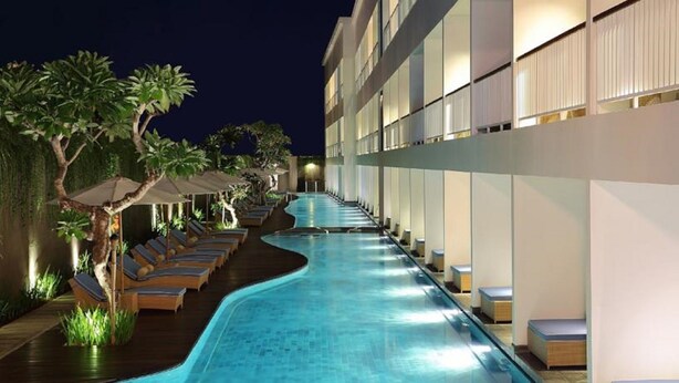 A swimming pool with lounge chairs and umbrellas, surrounded by a building with multiple floors. - Ossotel Legian Bali (Legian)