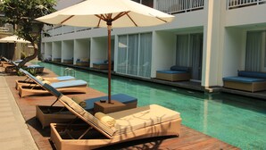 A poolside area with lounge chairs and an umbrella, a building with balconies, and a clear pool. - Ossotel Legian Bali (Legian)