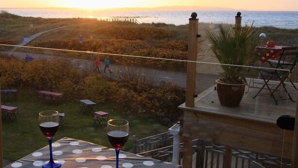 Un balcon avec vue sur le coucher de soleil sur la mer, deux verres de vin rouge posés sur une nappe à pois et une plante en pot.