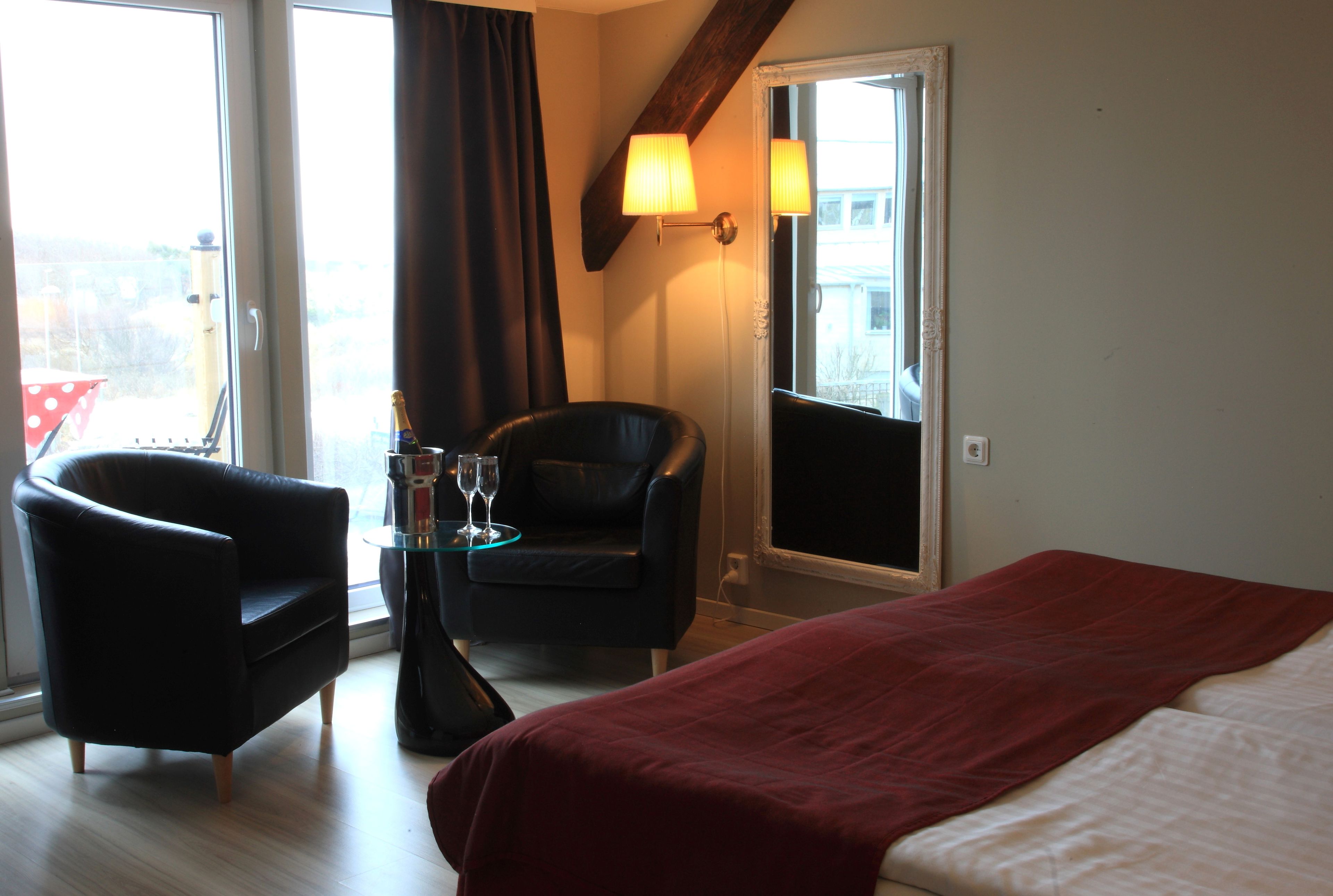 A hotel room with a bed, a chair, a small table with a bottle and glass, and a window with curtains.