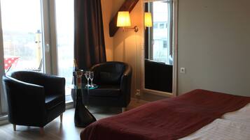 Habitación de hotel con cama, una silla, una mesita con una botella y un vaso, y una ventana con cortinas.