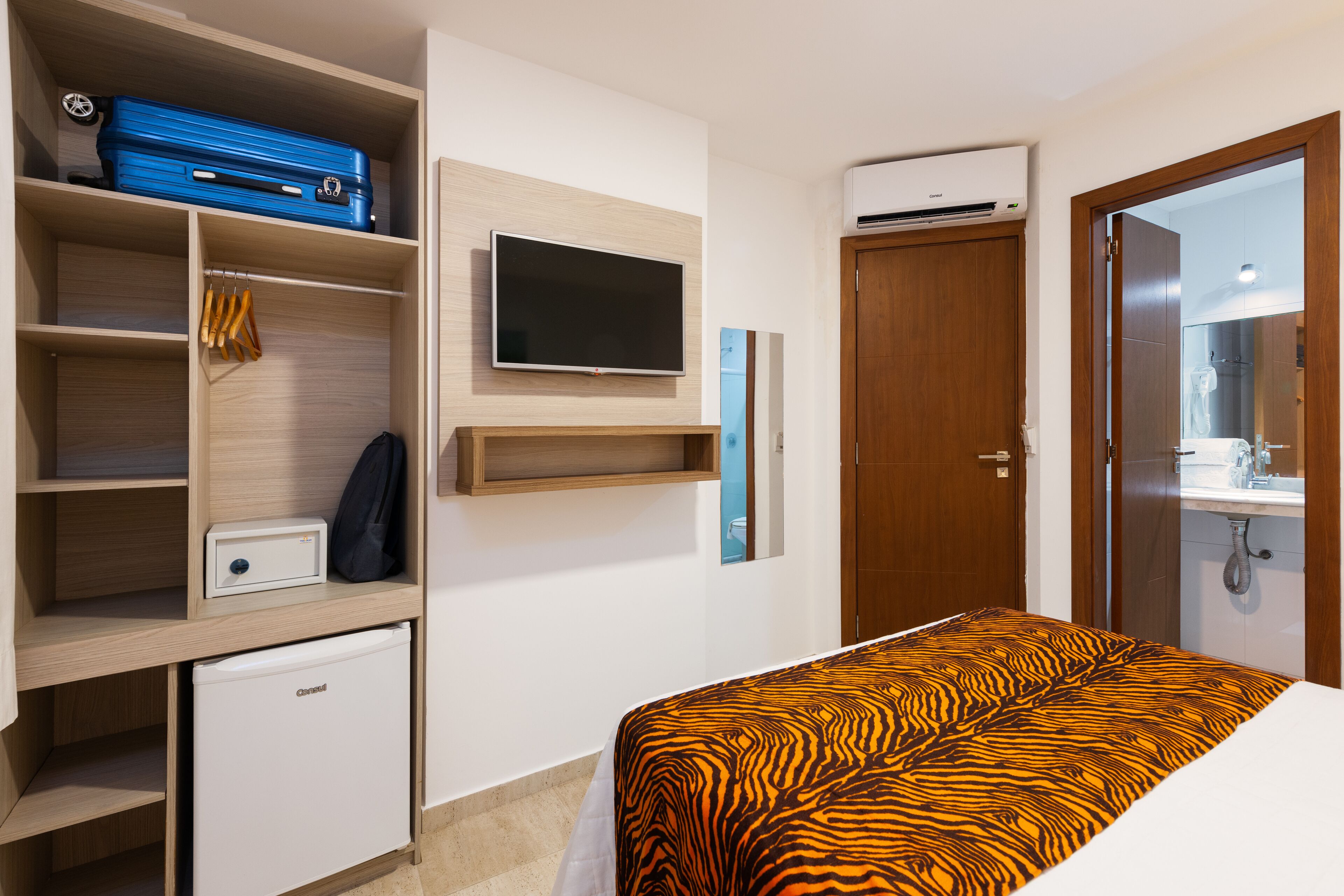 Habitación de hotel con cama, televisión, armario, refrigerador pequeño y baño visible a través de una puerta abierta.