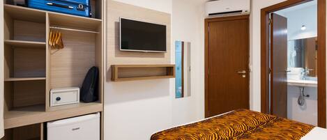 Habitación de hotel con cama, televisión, armario, refrigerador pequeño y baño visible a través de una puerta abierta.