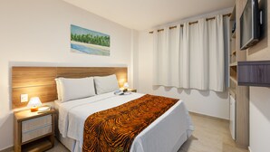 Una habitación de hotel con una cama, una mesita de noche con una lámpara, un televisor montado en la pared y un cuadro de una playa.