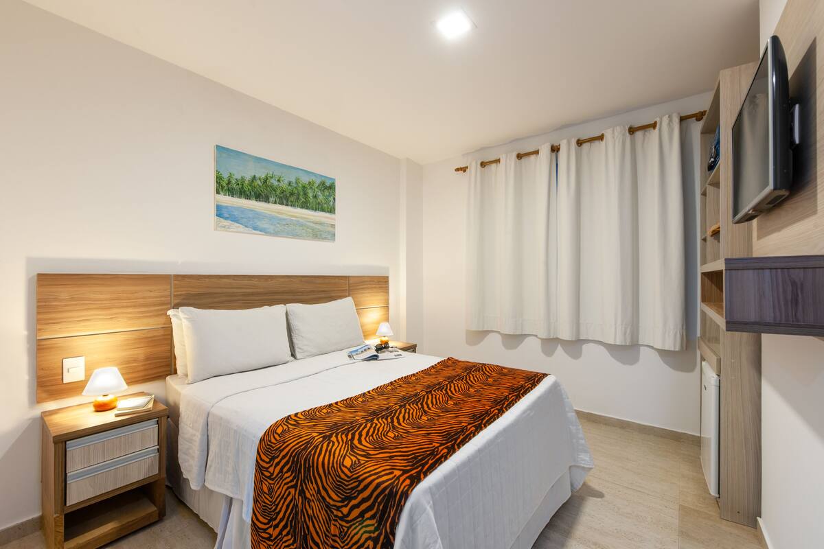Une chambre d’hôtel avec un lit, une table de chevet avec une lampe, un téléviseur fixé au mur et un tableau représentant une scène de plage.