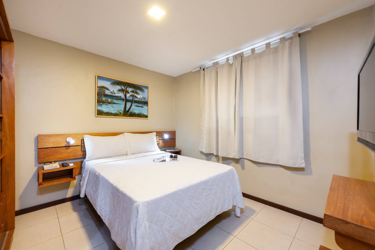 Une chambre d’hôtel avec un lit, une tête de lit en bois, des tables de chevet, un tableau représentant une scène de plage, une fenêtre avec des rideaux et un bureau en bois.
