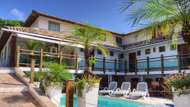 Un área junto a la piscina con sillones de descanso, una palmera y un edificio con techo de tejas.