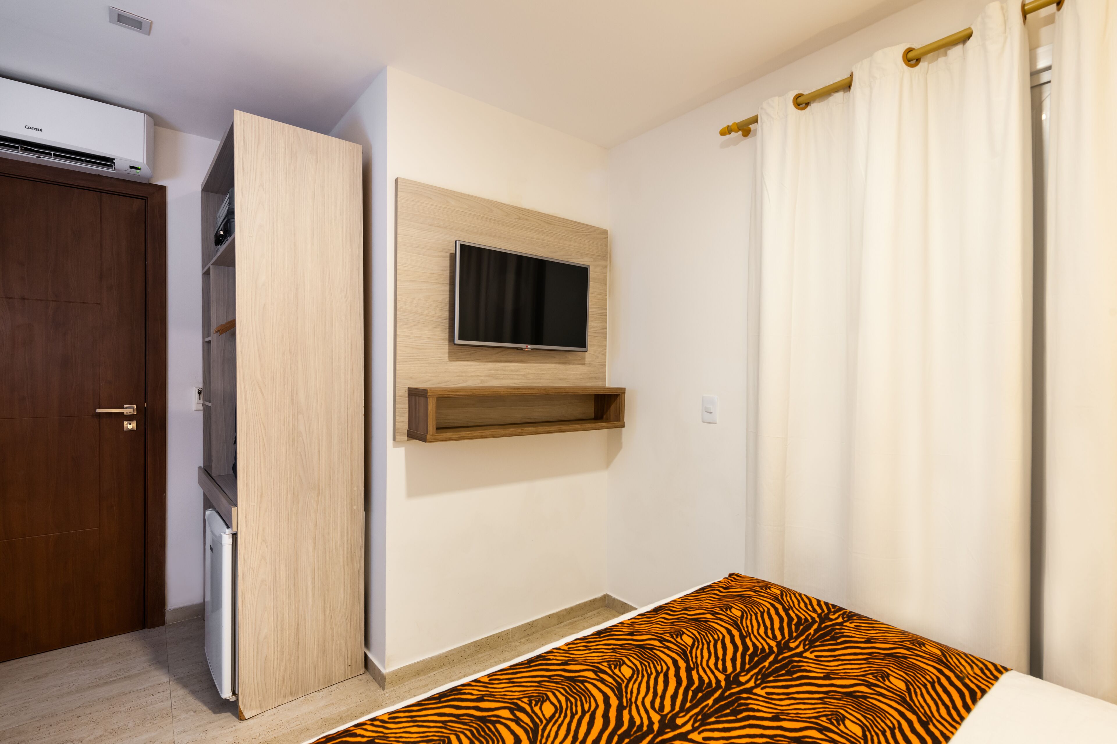 A bedroom with a bed, a mounted TV, a built-in shelf, a wardrobe, and a white curtain.