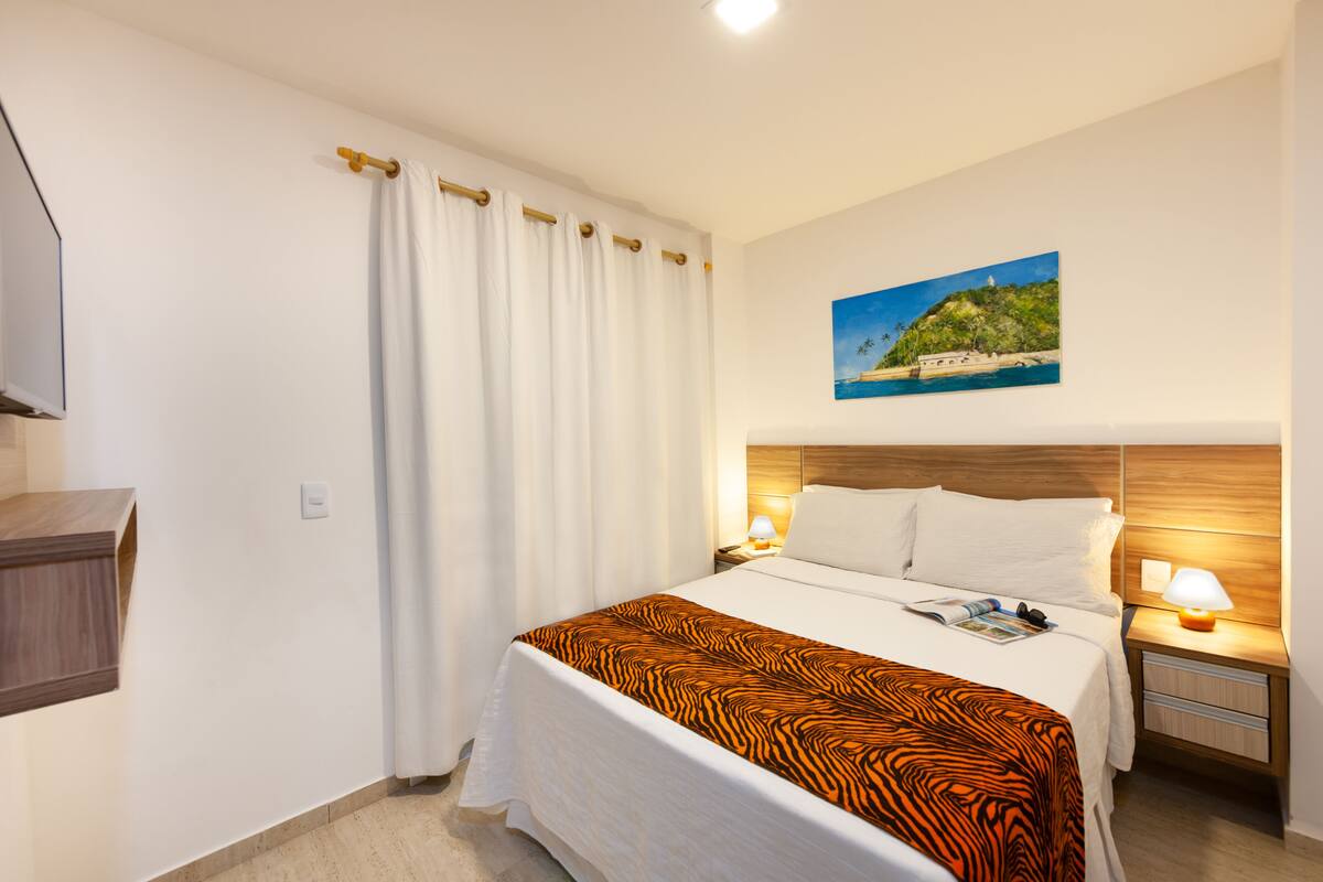 Une chambre d’hôtel avec un lit, une table de chevet avec une lampe, un tableau au mur et une fenêtre avec des rideaux.