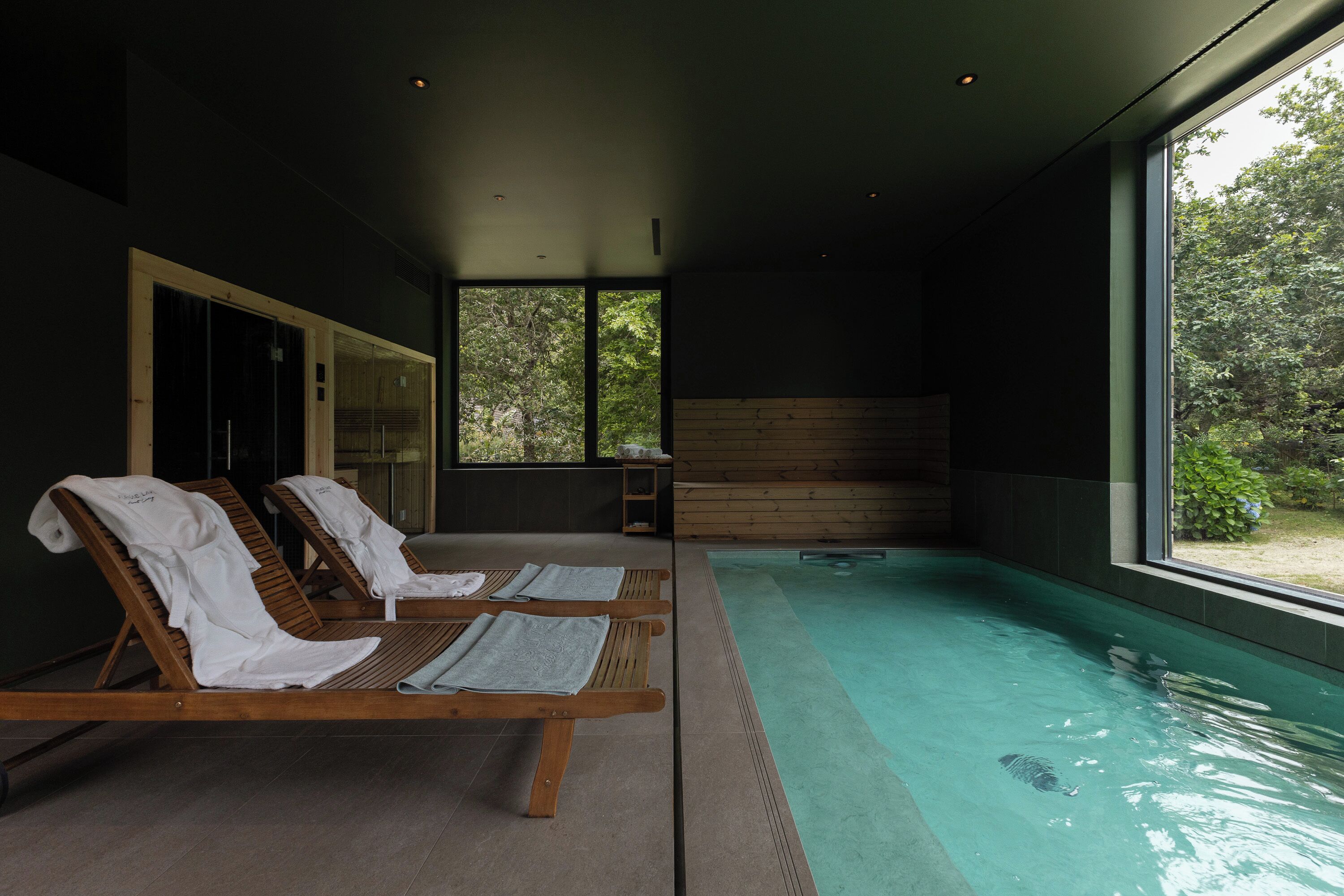 A modern indoor swimming pool with wooden loungers and a glass wall.