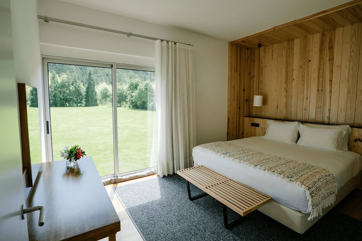Een slaapkamer met een groot bed, een houten hoofdbord, een bankje en uitzicht op een groen gazon door een schuifdeur.