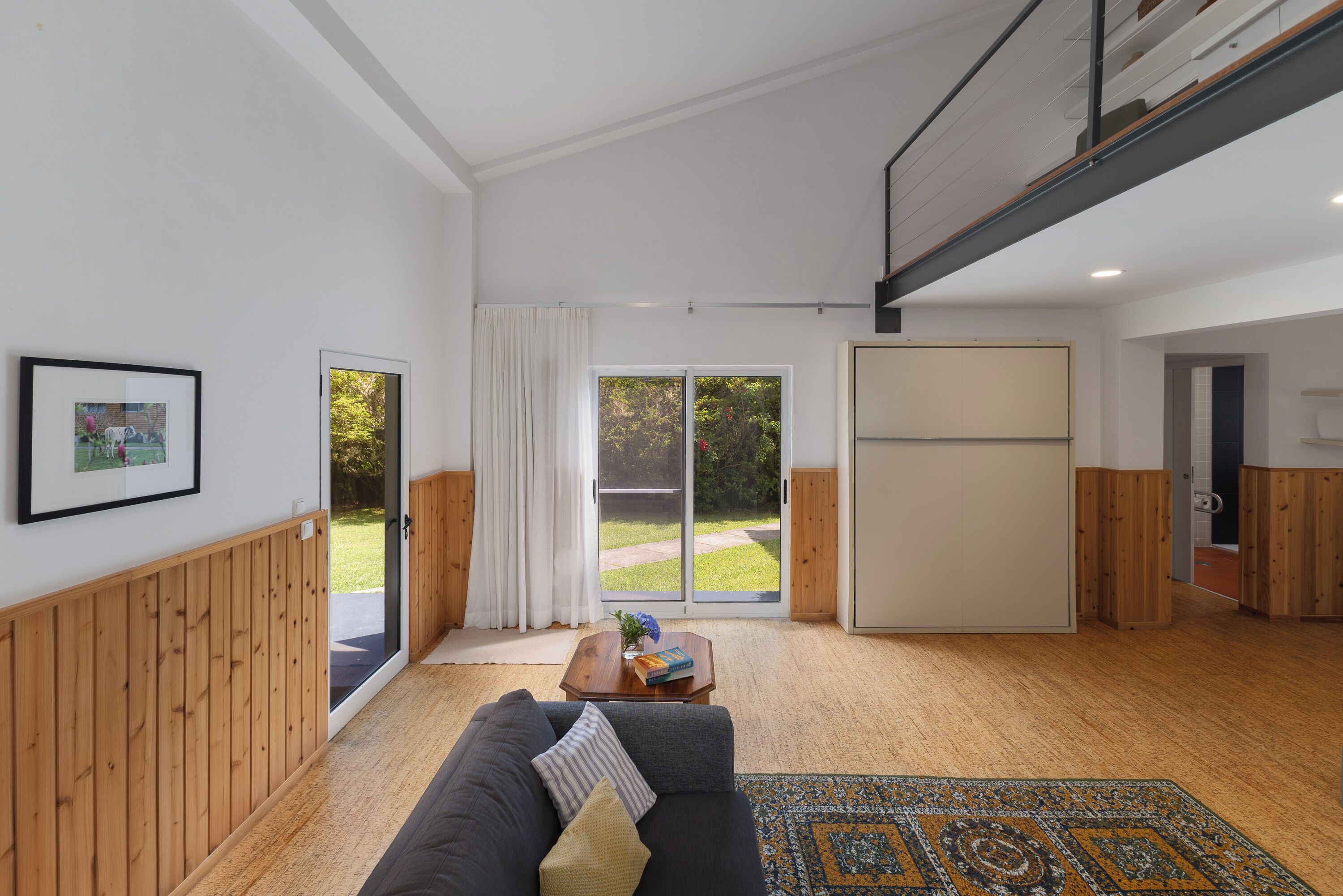 A modern living room with a sofa, a coffee table, and a patterned rug. There is a sliding glass door leading to a garden.