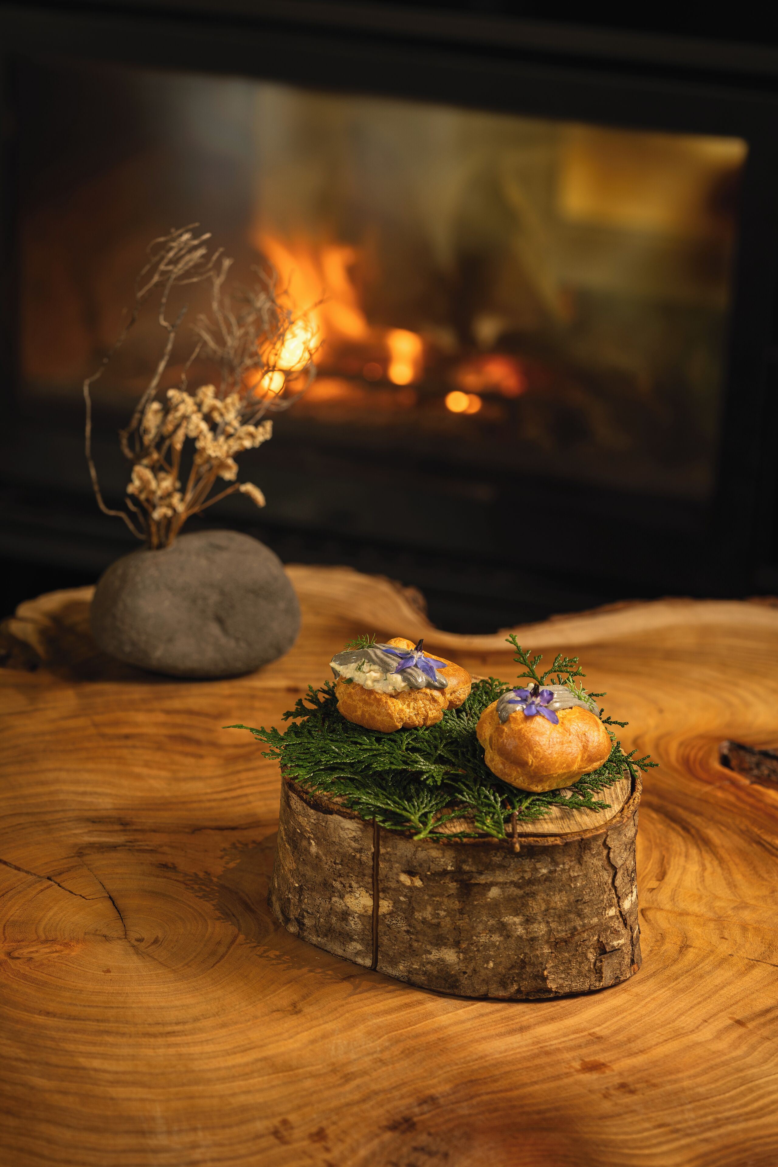 A rustic wooden table with two small pastries on a wooden log centerpiece, set against a backdrop of a fireplace with a warm glow.