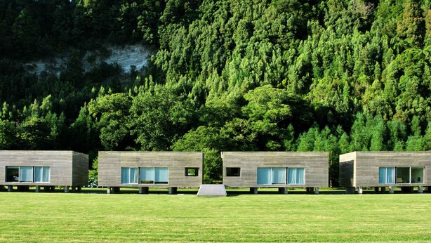 Tres cabañas modernas con ventanas amplias, elevadas sobre pilares en un campo cubierto de hierba, con un bosque denso al fondo.