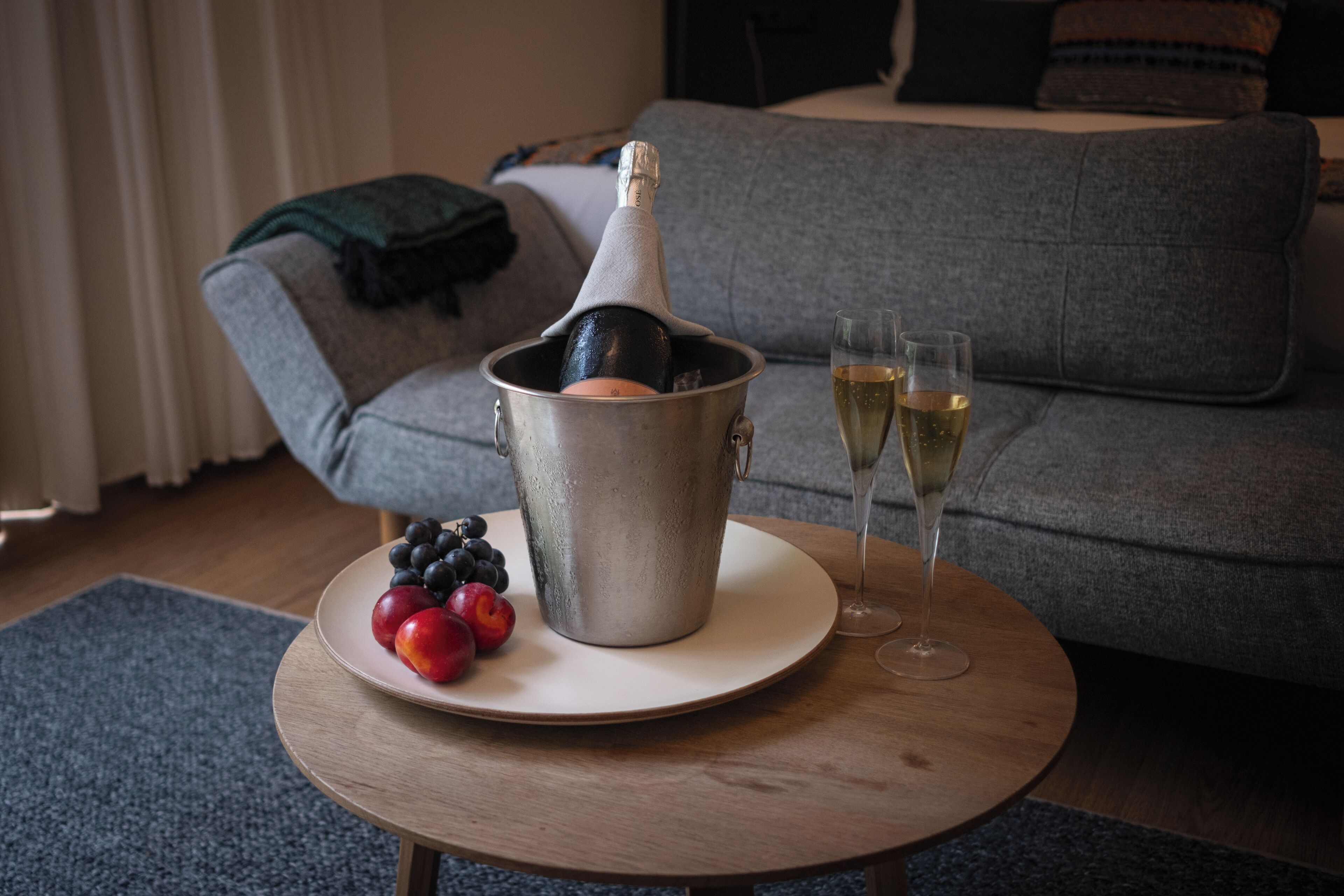 Une pièce avec un canapé gris, une table basse en bois sur laquelle se trouve une bouteille de champagne, deux verres à champagne et une assiette de fruits.