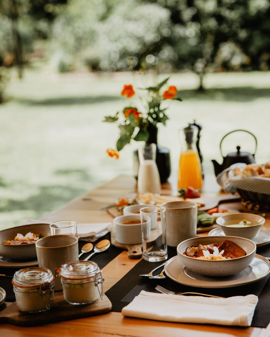 Una mesa de desayuno al aire libre con diferentes platos, vasos y un jarrón de flores.