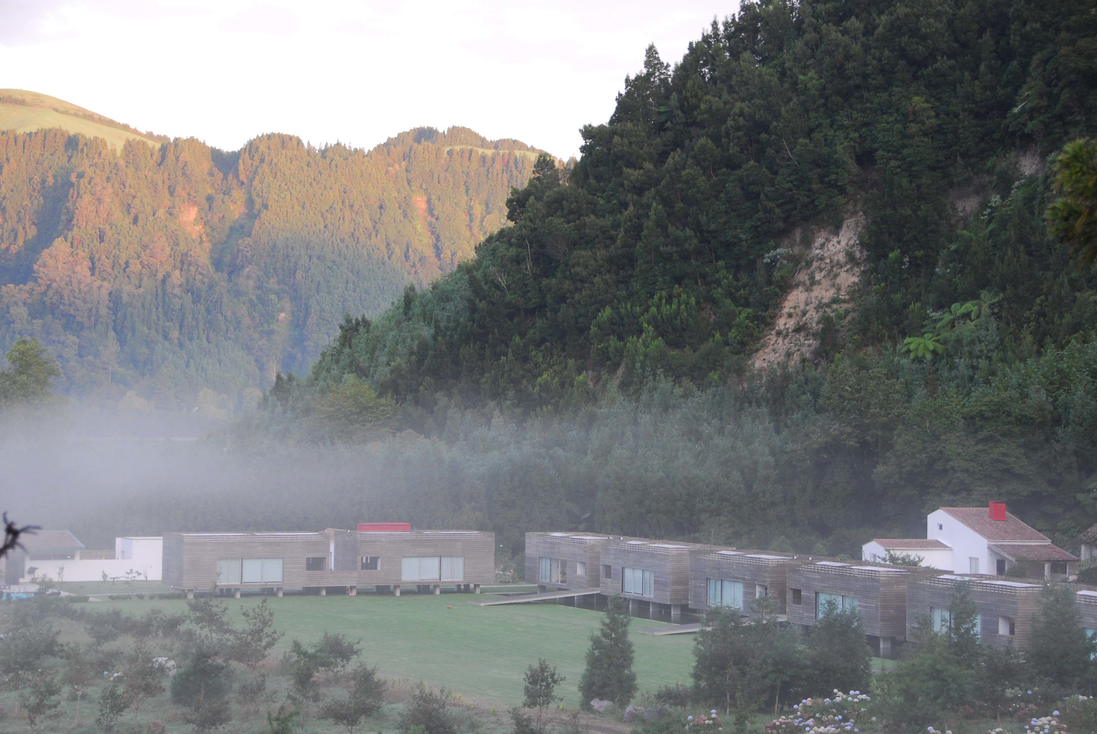 A row of modern, multi-story buildings with large windows, set against a backdrop of a forested hillside.