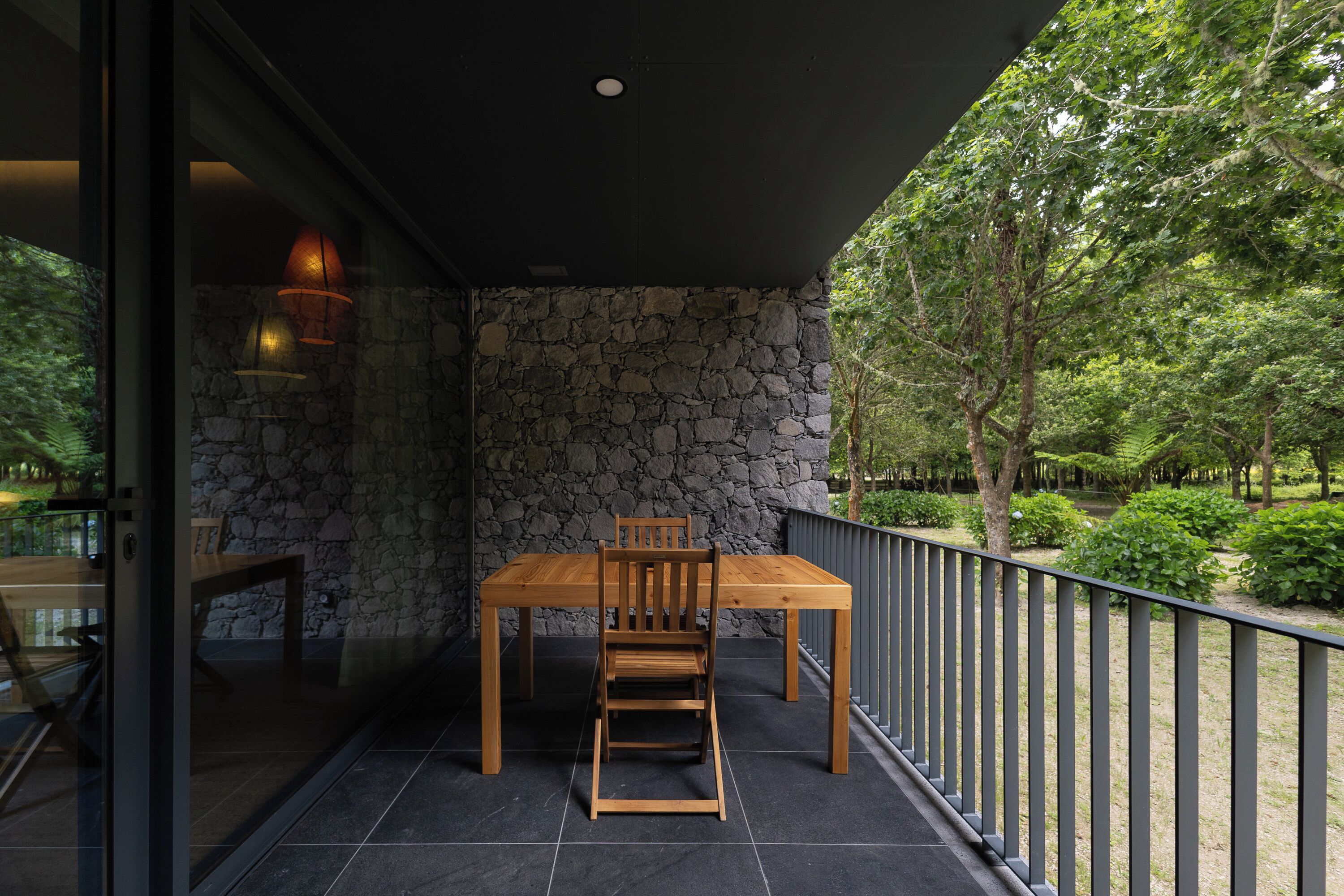 A balcony with a stone wall, a wooden table and chairs, and a view of a garden.
