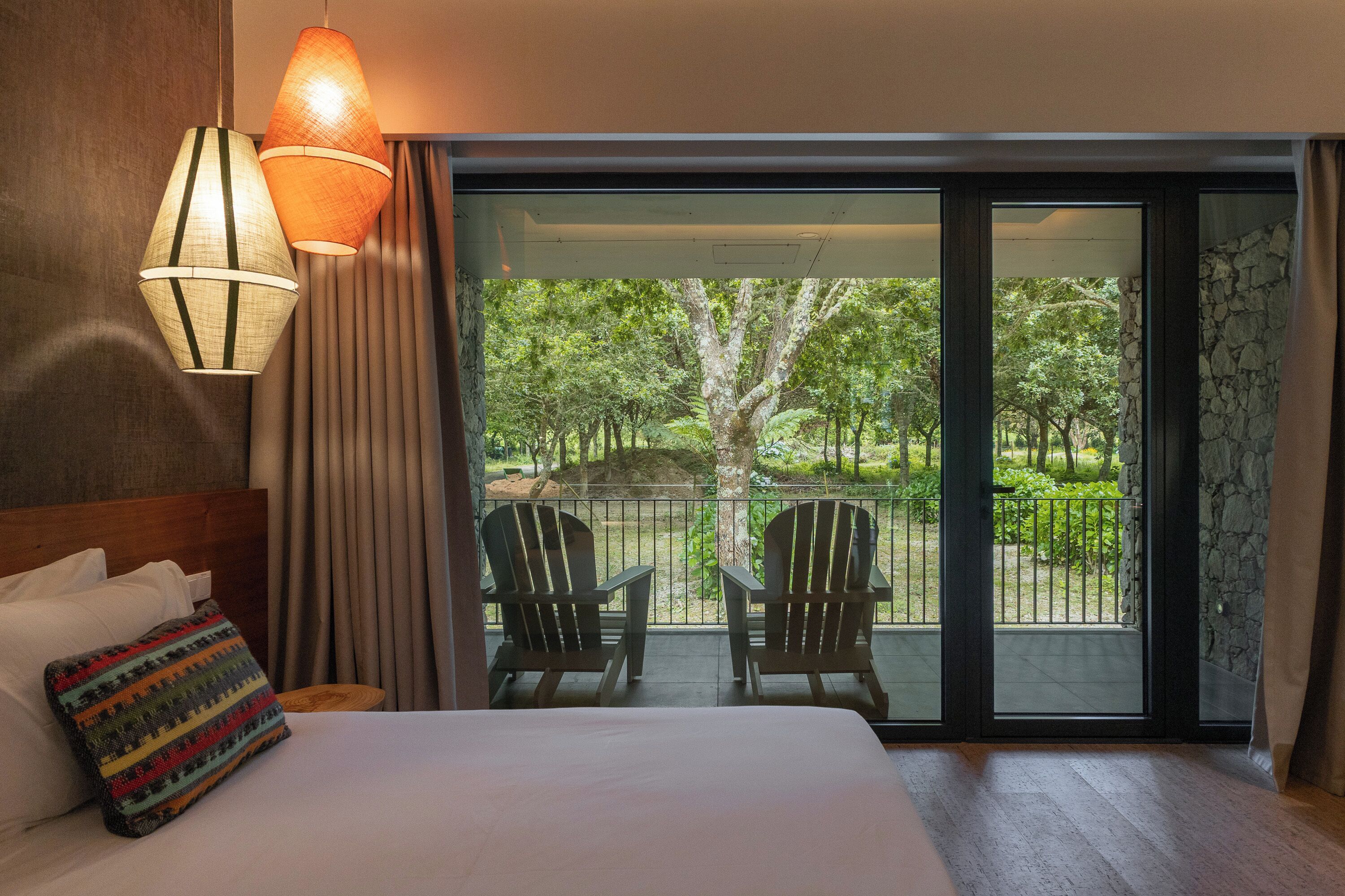 A hotel room with a bed, two chairs, a view of a garden through a sliding door, and a hanging lamp.
