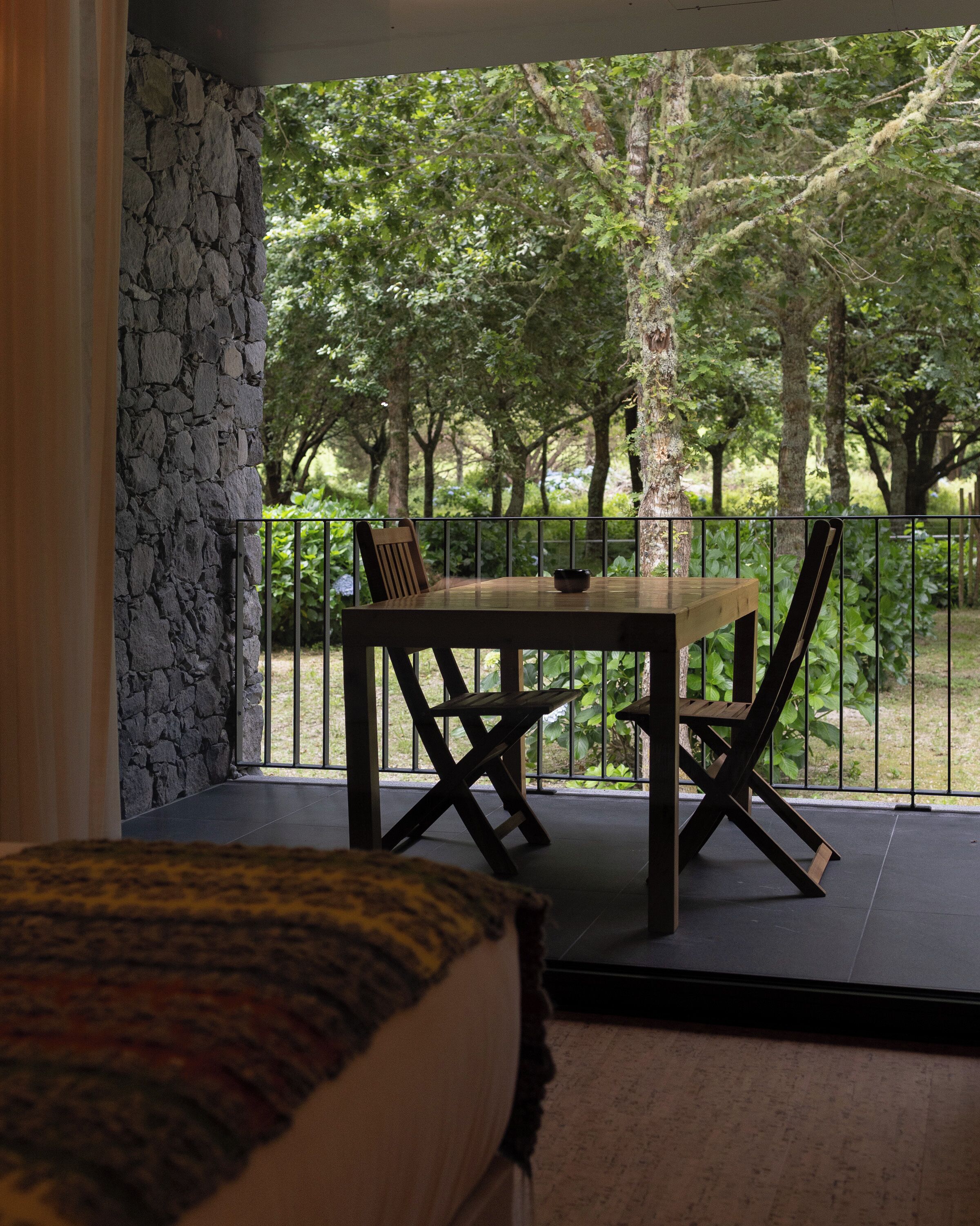 A balcony with a stone wall, a table with chairs, and a view of a forested area.