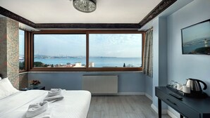 A hotel room with a large window offering a view of the sea and city skyline, a bed with white linens, a radiator, and a small table with a kettle and cups. - Mytra Hotel (Istanbul)