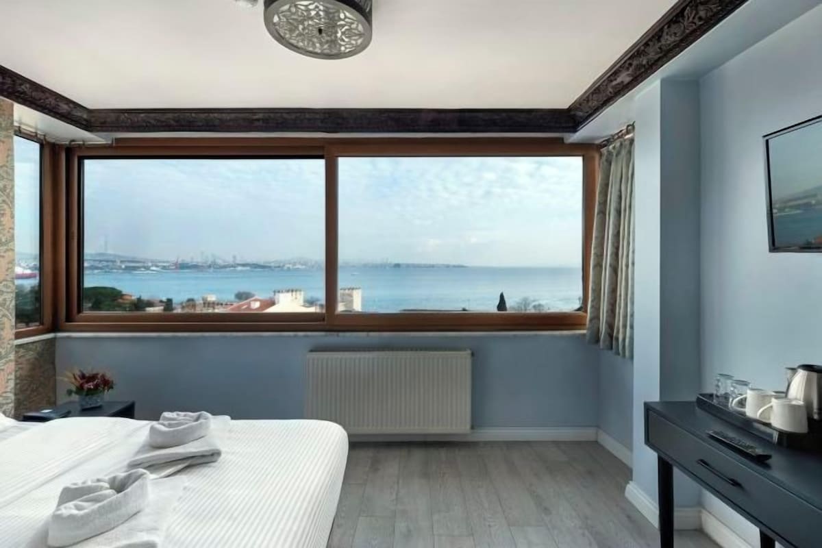 Un cuarto de hotel con una ventana grande, una cama, un radiador, un escritorio y vista al mar.