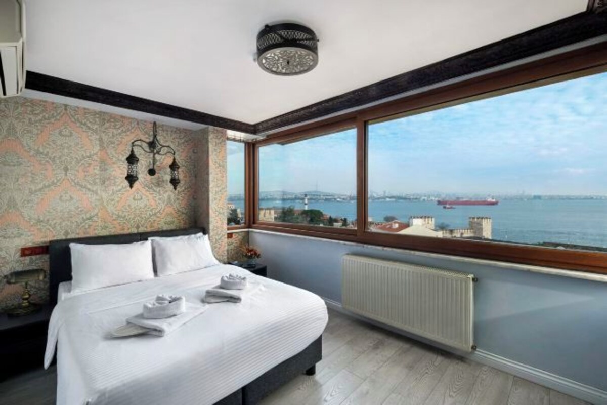 A hotel room with a large window, a bed, a radiator, and a wall-mounted light fixture.