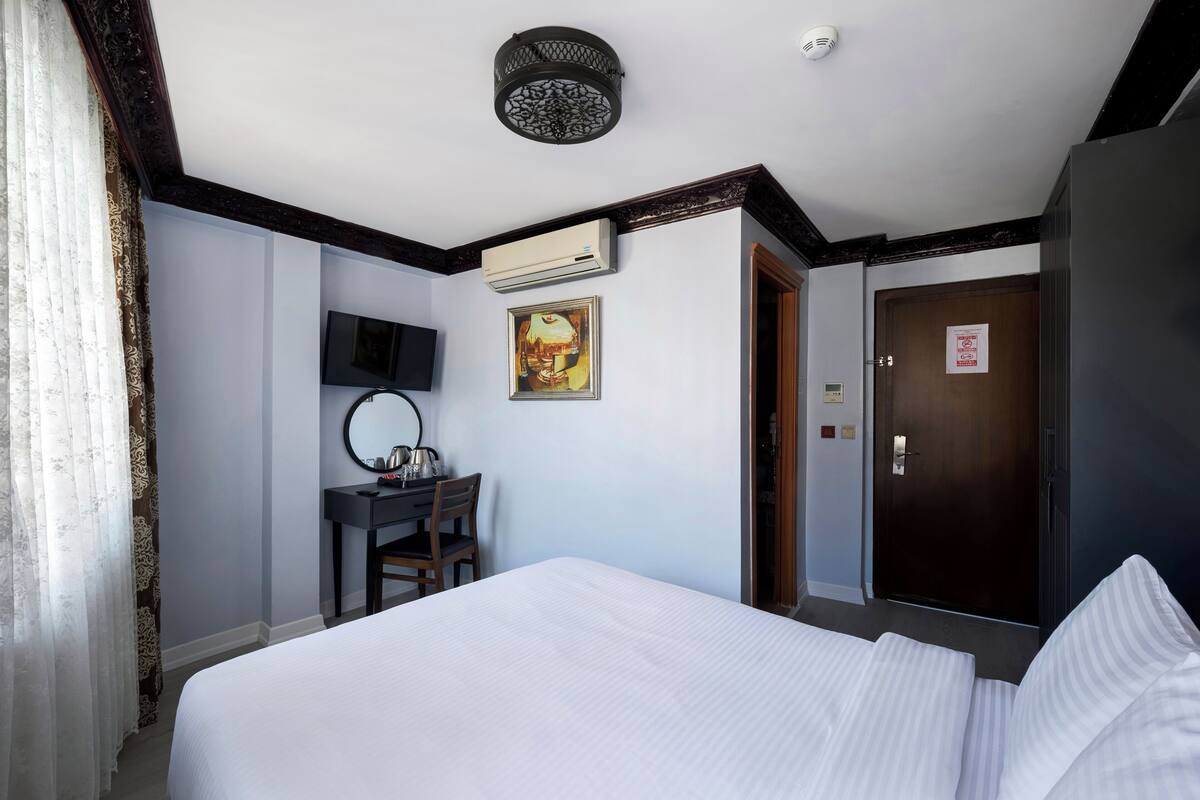A hotel room with a bed, a desk with a chair, a flat-screen TV, an air conditioning unit, a door with a sign, and a ceiling light fixture.
