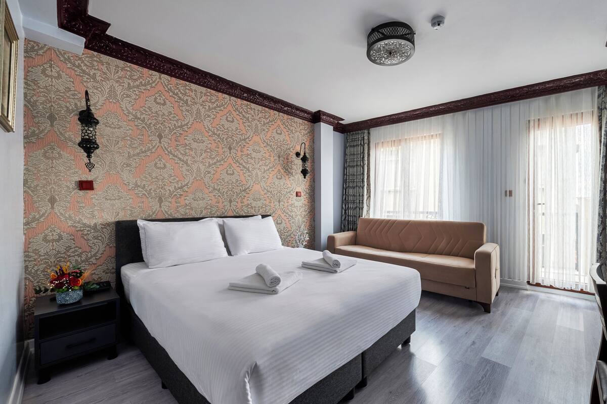 A hotel room with a large bed, a sofa, a desk with a computer, and a window with curtains.
