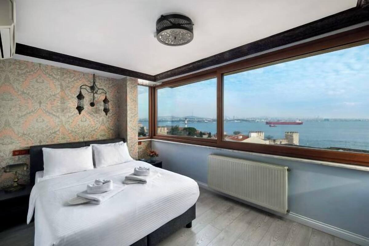 A hotel room with a large window, a bed, a radiator, and a wall-mounted light fixture.
