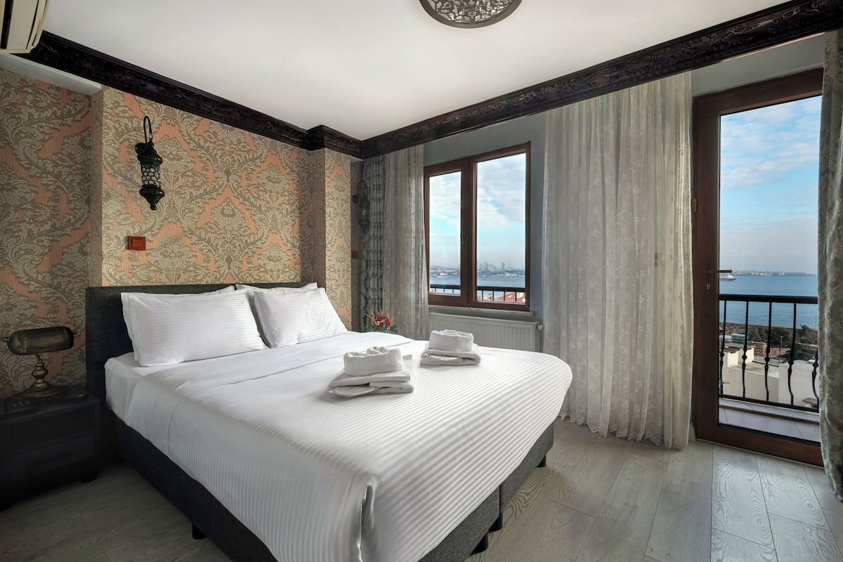 A hotel room with a large bed, a view of the city through the window, and a balcony with a railing.