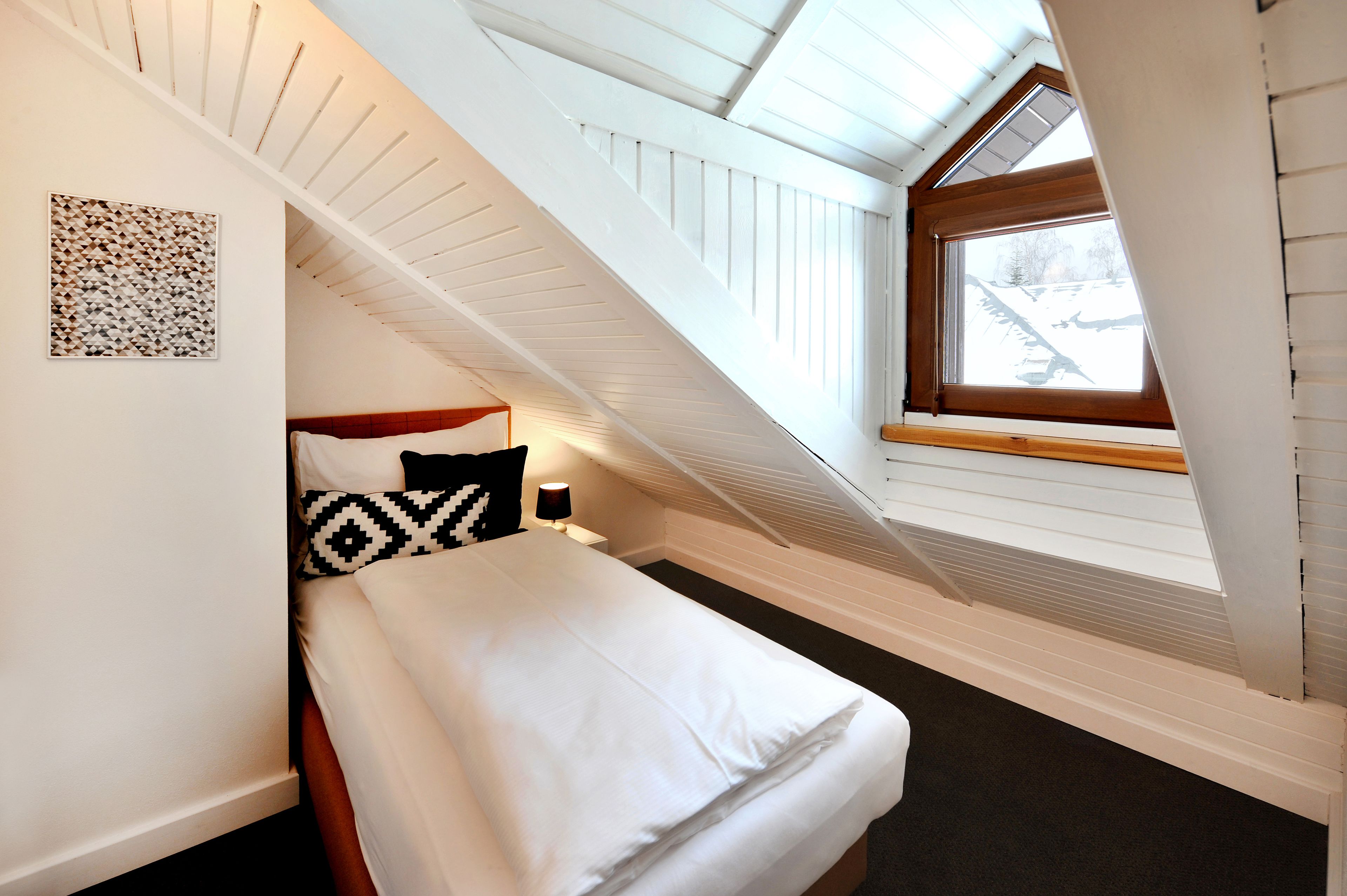 A small, cozy bedroom with a sloped ceiling, a single bed, a nightstand, a wall-mounted lamp, and a window with a view of the outside.