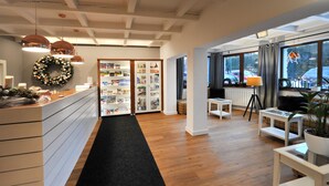 Un área de recepción con un mostrador de madera, alfombra negra, una pared con una corona decorativa, una vitrina de vidrio con revistas y una ventana con vistas a coches estacionados.
