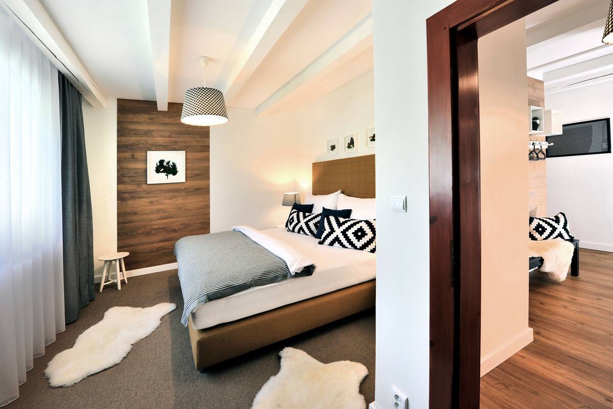 Quarto moderno com cabeceira de madeira, uma cama grande com almofadas estampadas, um banquinho pequeno e um tapete de pele no chão.