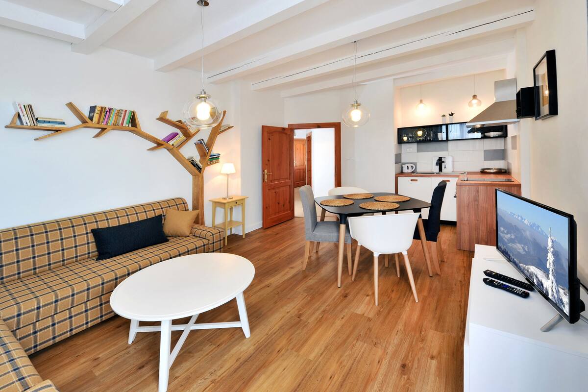 Uma sala de estar moderna com um sofá, uma mesa redonda e um espaço de refeições com cadeiras. É possível ver uma cozinha ao fundo, com uma televisão e controles remotos sobre uma mesa.