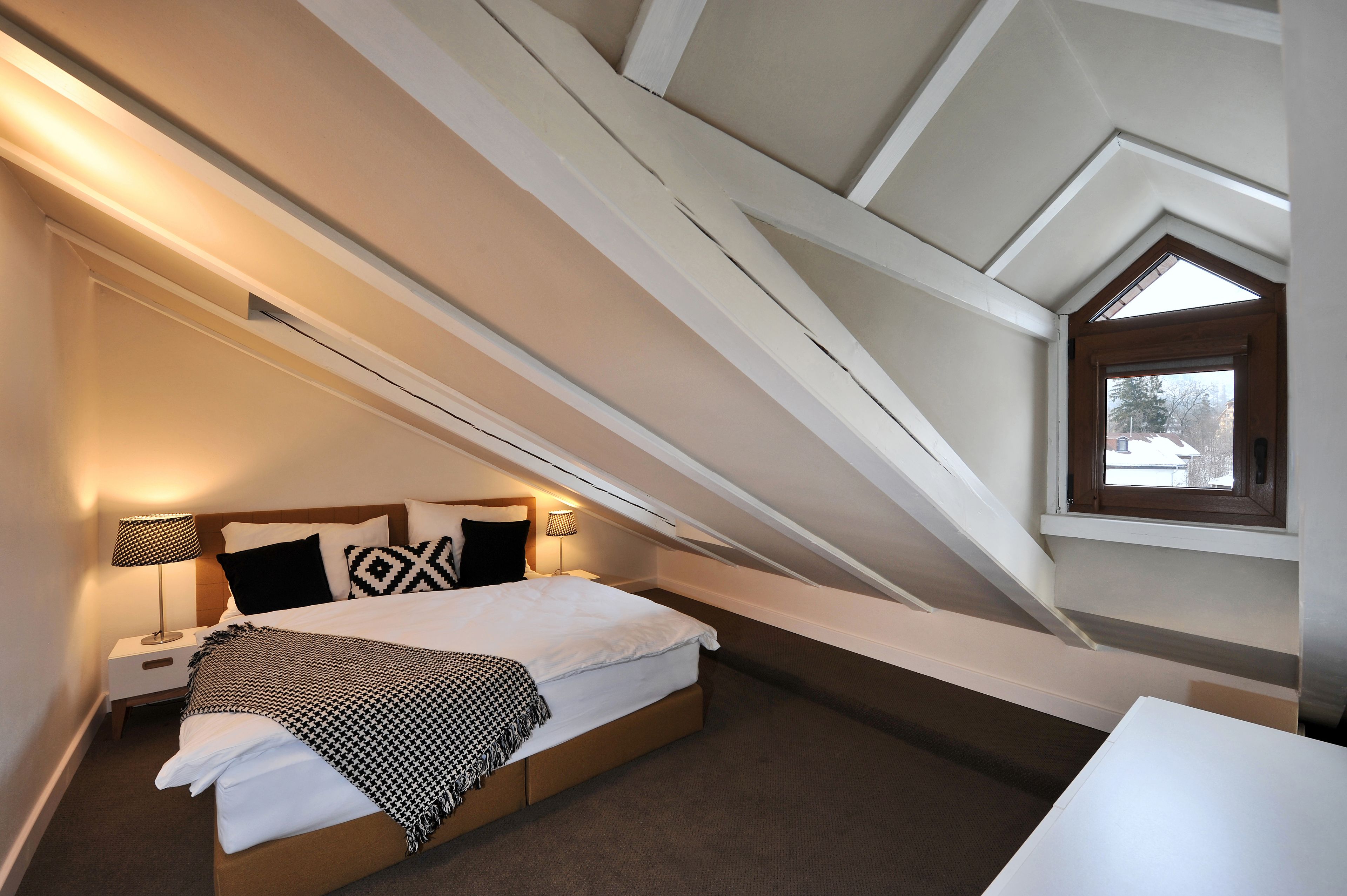 Een slaapkamer met een schuine plafond, een bed met witte lakens, een nachtkastje met een lamp, en een raam met uitzicht op bomen.