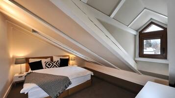 Een slaapkamer met een schuine plafond, een bed met witte lakens, een nachtkastje met een lamp, en een raam met uitzicht op bomen.