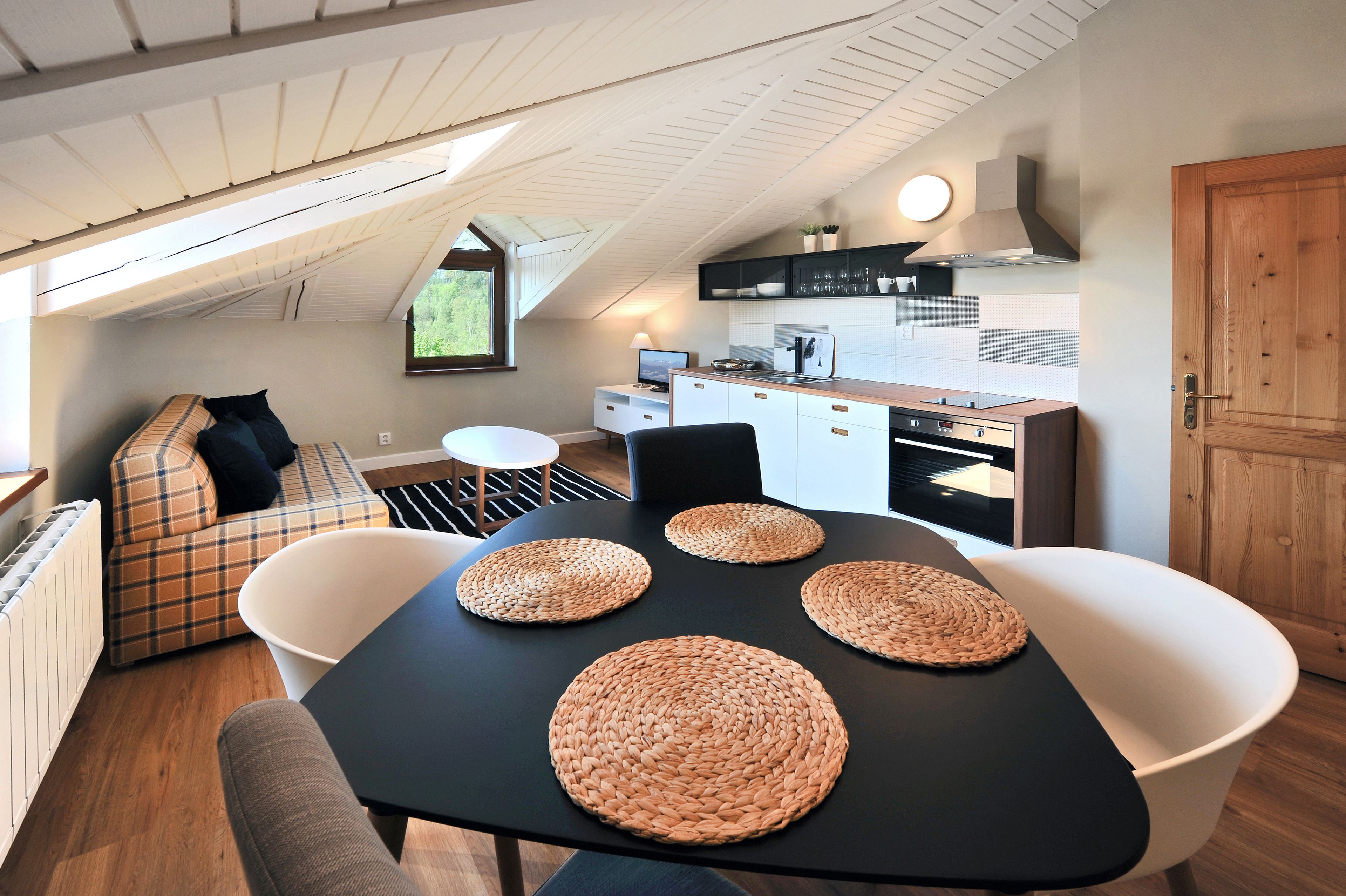 A cozy loft-style room with a dining area featuring a black table and woven placemats, a sofa with plaid cushions, and a kitchen with modern appliances and a wooden door.