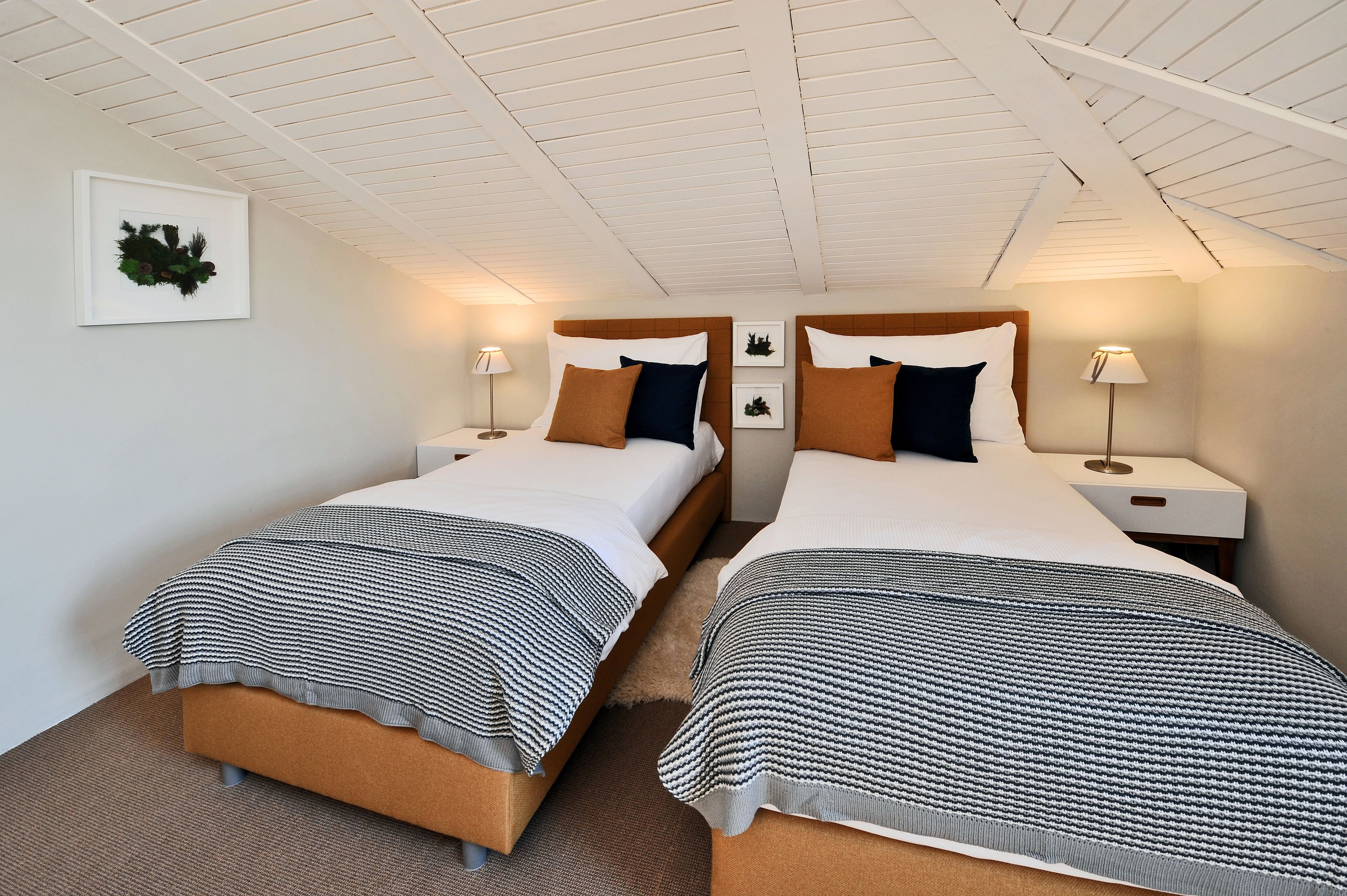 Habitación de hotel con dos camas, cada una con cabecero, mesitas de noche y lámparas. La habitación tiene techo inclinado y un cuadro enmarcado en la pared.
