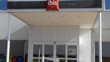 Entrada de un hotel con un área techada, puertas de vidrio y un letrero rojo de 'ibis'.
