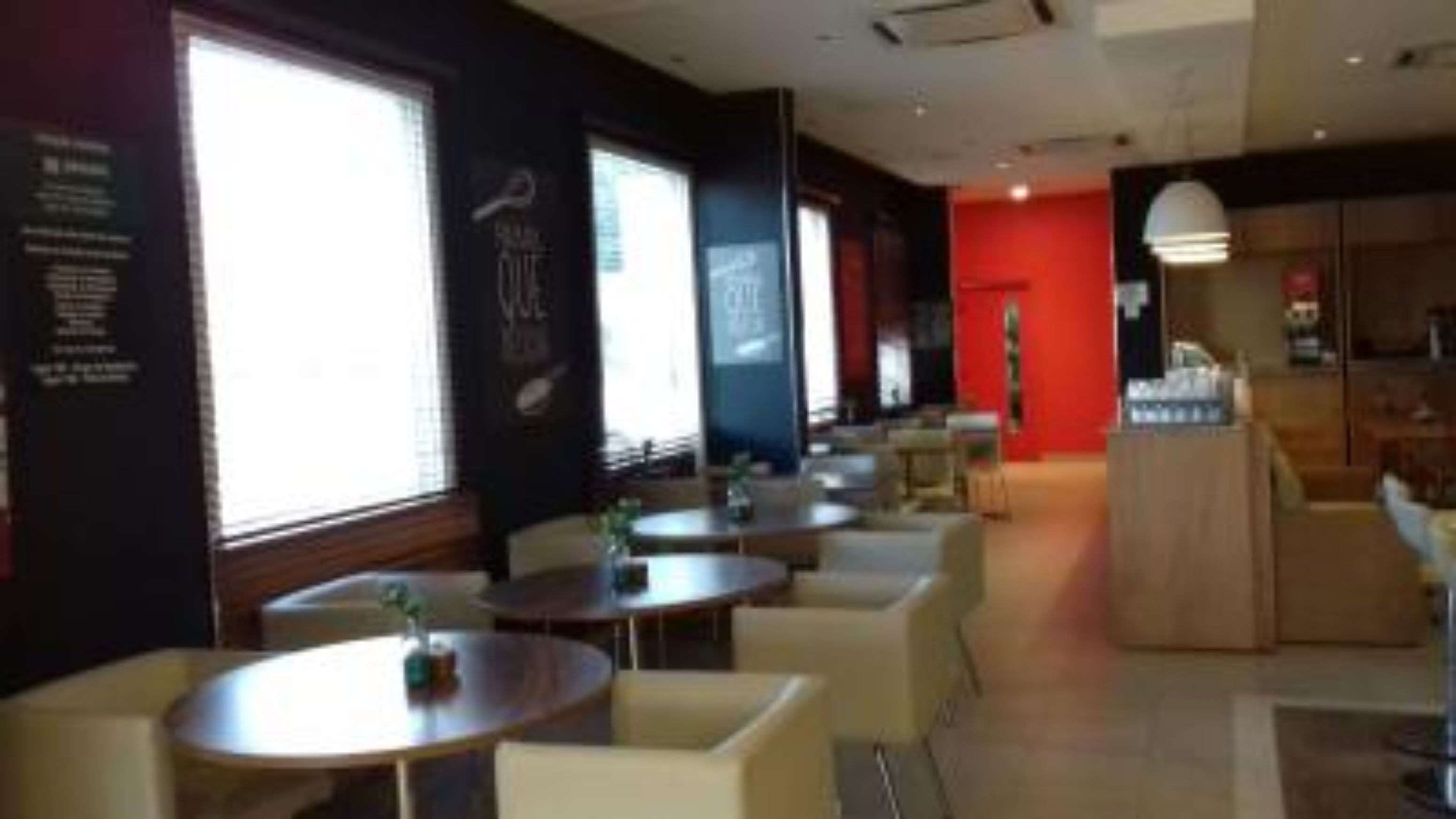 Interior moderno de restaurante con mesas y sillas redondas, ventanas amplias y una pared de acento roja.