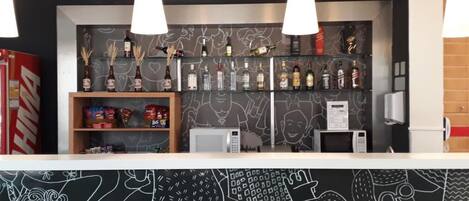 Zona de bar con un mural en blanco y negro, estantes con botellas, aperitivos y un microondas.