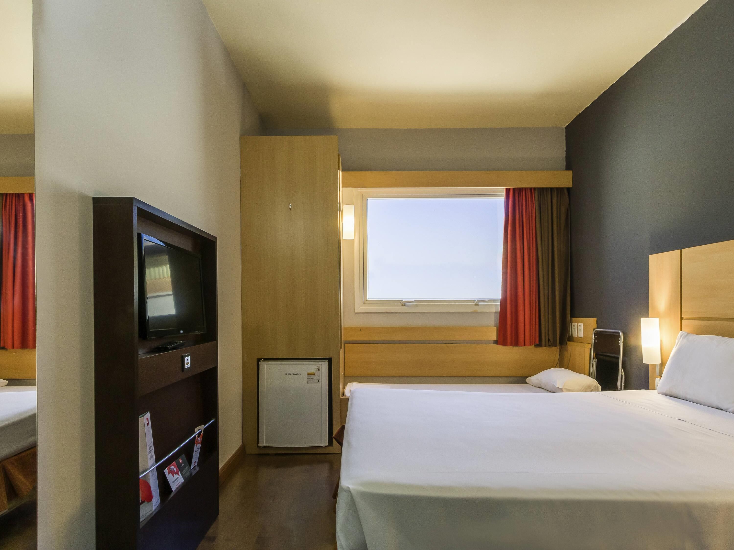 Quarto de hotel com cama, televisão, minibar, janela com cortinas e mesa de cabeceira com abajur.