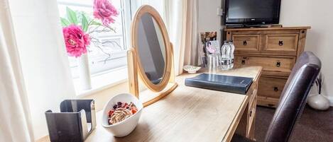 A hotel room with a wooden desk, a mirror, a television on a wooden dresser, and a window with a view of flowers.
