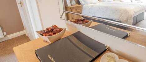 A hotel room with a wooden desk, a bowl of snacks, a guest information booklet, and a bed with white bedding.