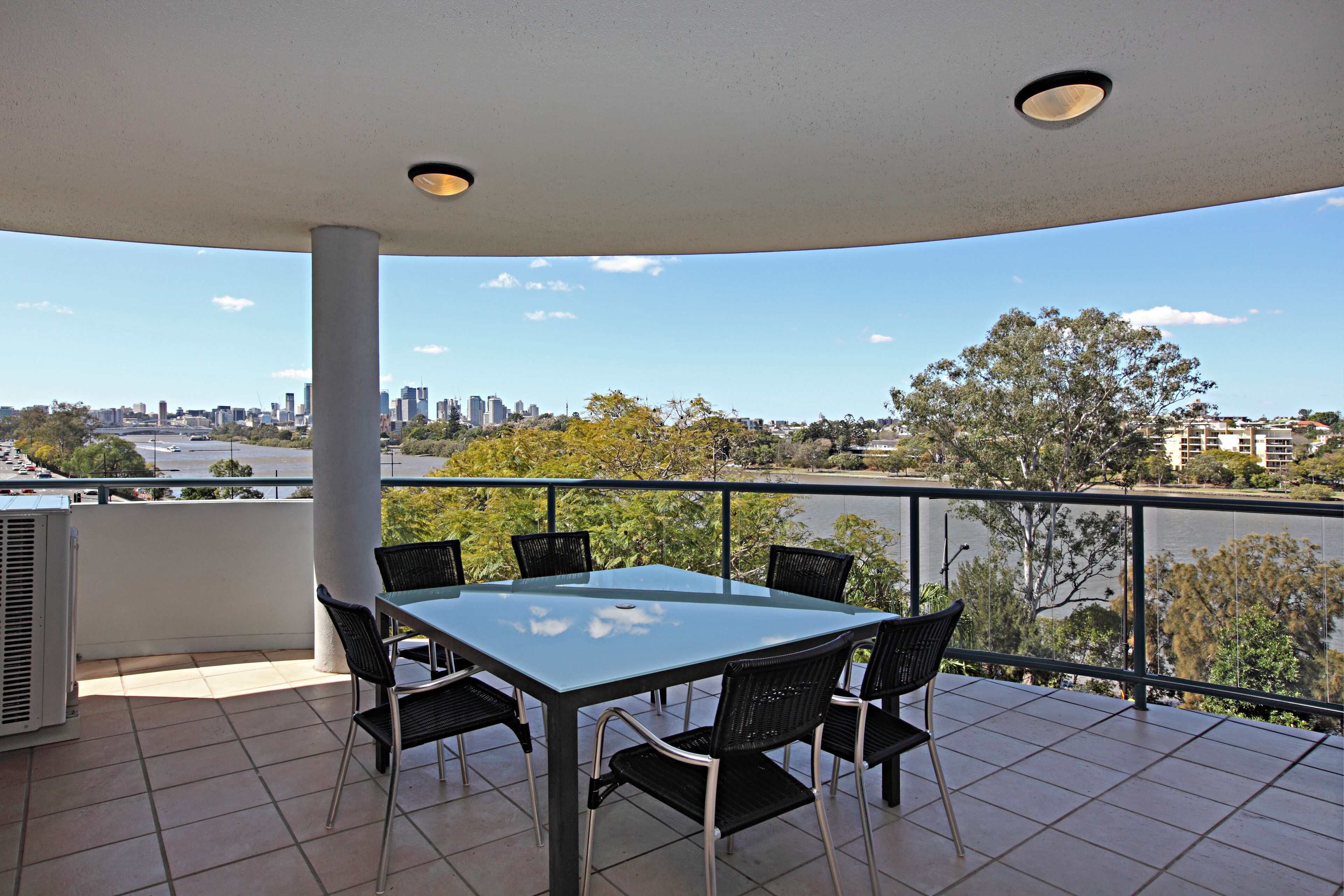 A balcony with a glass table and chairs, overlooking a river and city skyline.