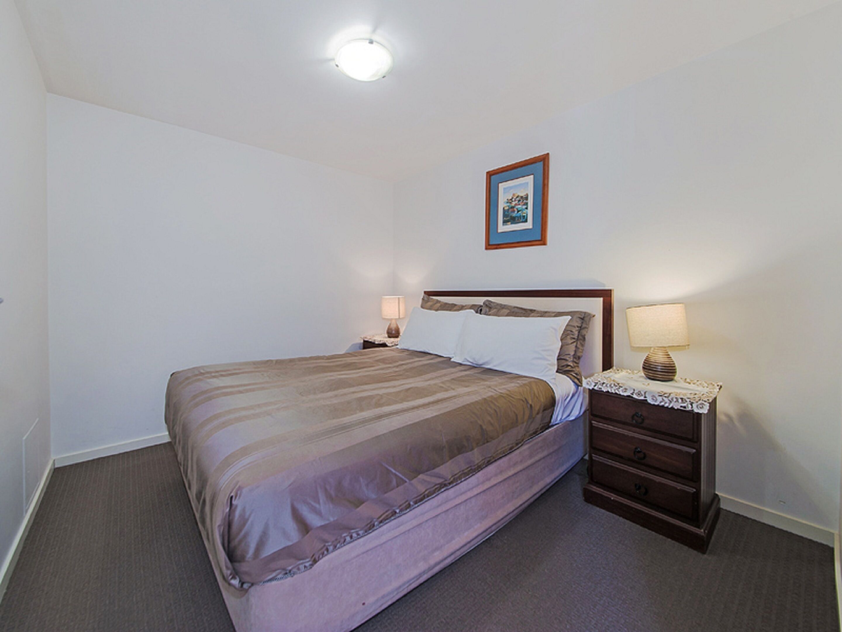 Ein ordentlich gemachtes Bett mit gestreifter Decke, ein Nachttisch mit Lampe und ein gerahmtes Bild an der Wand.
