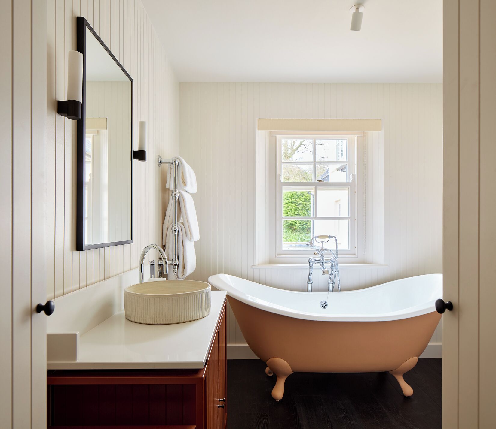 A bathroom with a freestanding bathtub, a round sink, a mirror, and a window with a view of greenery.