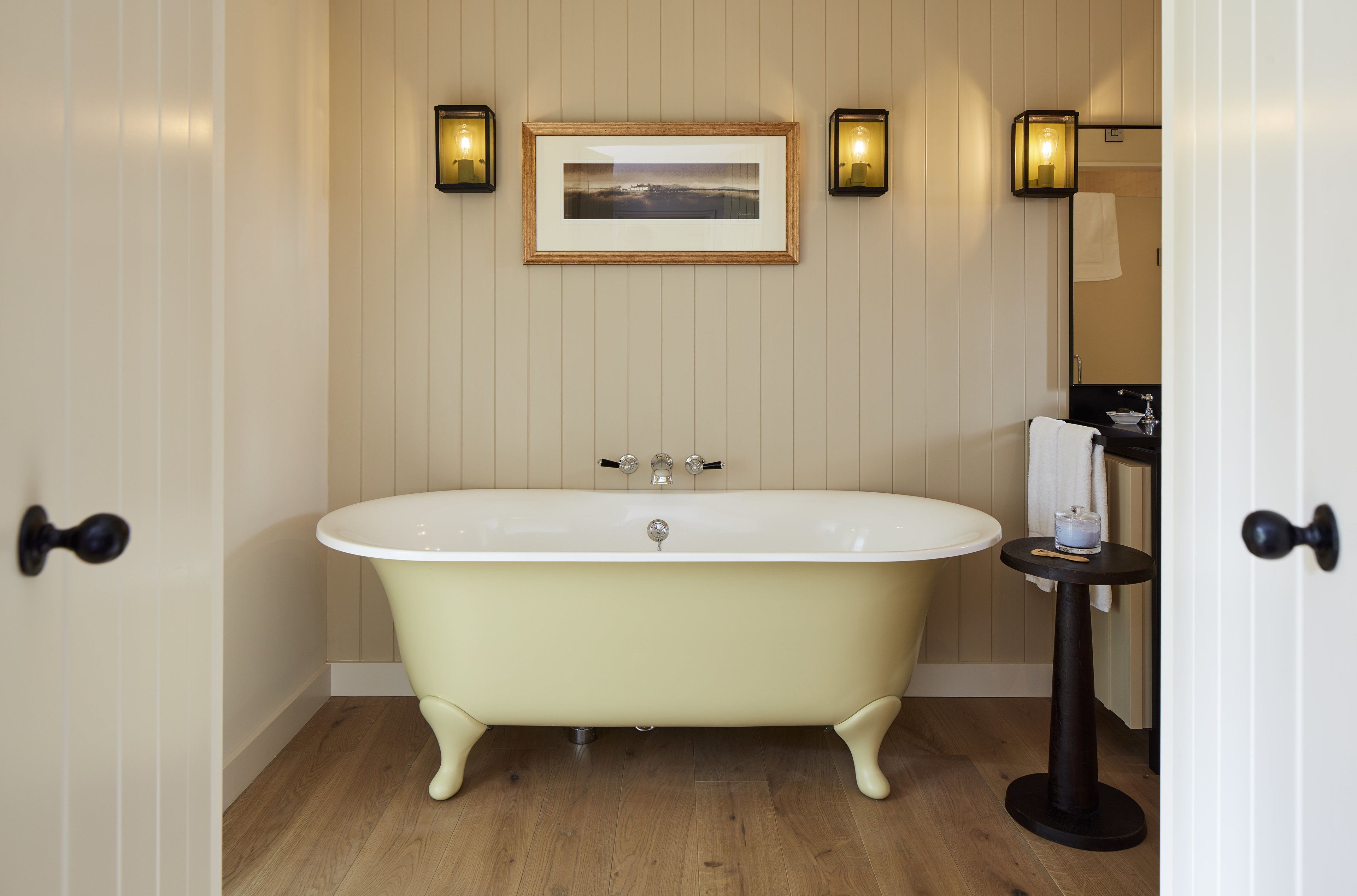 A vintage clawfoot bathtub in a bathroom with wooden paneling, a framed picture above the tub, and a small table with a candle.