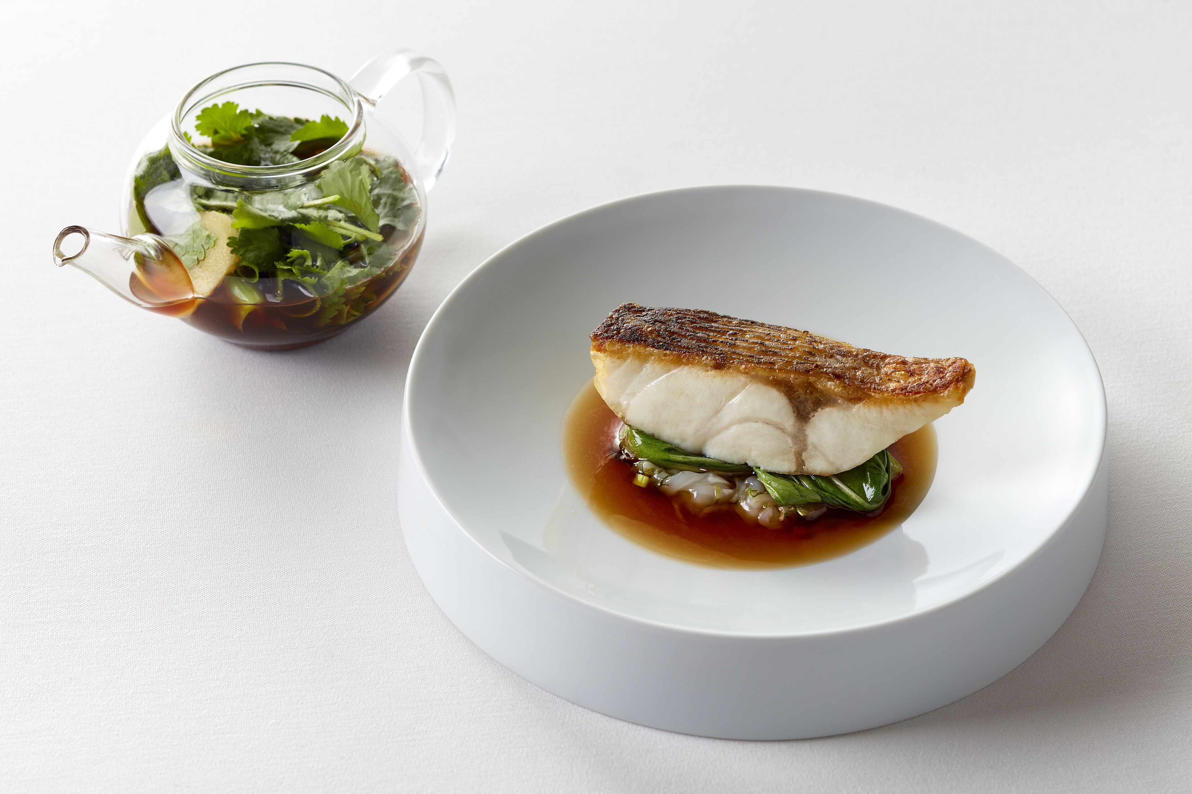 Un plato blanco con un trozo de pescado a la parrilla y vegetales, y una jarra de vidrio con líquido oscuro y vegetales.